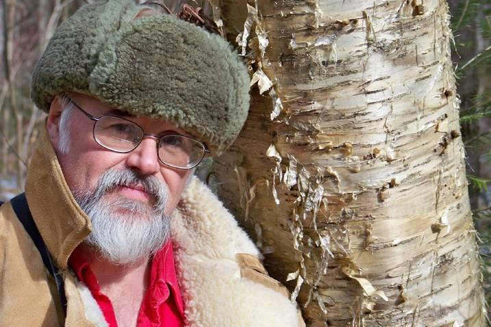 artist rick hunt leans against a golden peeling birch tree in big green winter wool hat and heavy jacket